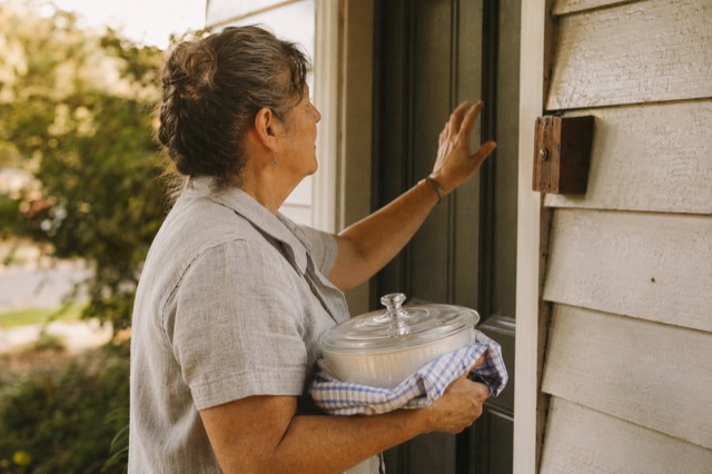 A middle-aged woman on a front porch carrying a covered casserole dish, lifting one hand toward the doorbell, afternoon light.