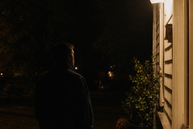 A single figure at the edge of a porch light's reach at night — calm and watchful, the moment before anything happens.