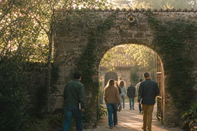 A retreat-center courtyard at dawn — an inconspicuous brass speaker mounted high on a stone wall above an arched gate, ivy growing up the wall.