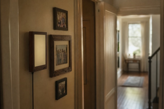 The walnut-framed e-ink Lamp mounted in a dim hallway beside small family photos, soft daylight.