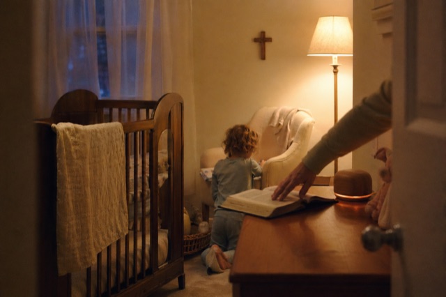 Bedtime in the nursery: a parent's hand visible setting an open children's Bible on the dresser, walnut Nursery device softly glowing, twilight blue through curtains.