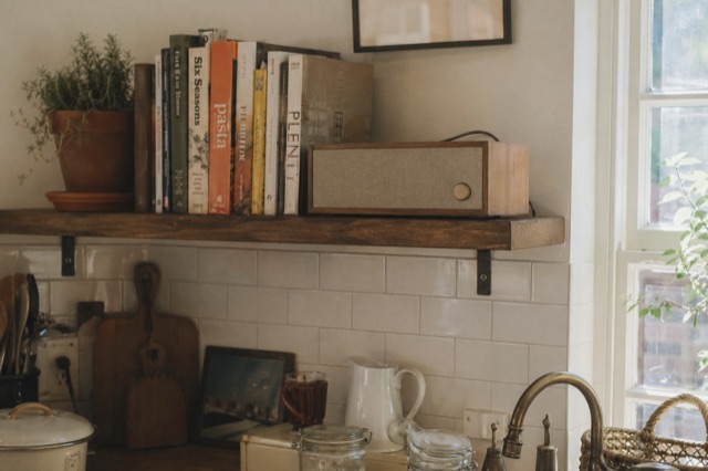 The walnut Prophet device on a high open shelf in a kitchen, well-used cookbooks, herb in a terracotta pot.