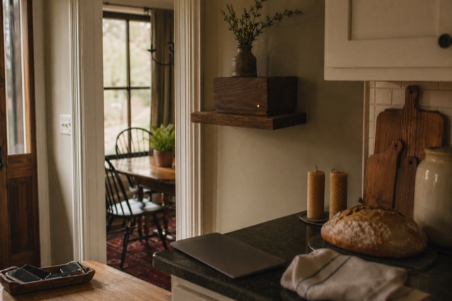 A family kitchen in late Friday afternoon golden light: bread cooling, unlit beeswax candles, phones face-down in a wooden tray, walnut device dimmed amber.