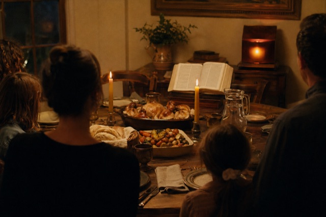 A Saturday-evening table with two lit beeswax candles, roast meal in pottery dishes, an open Bible at the head of the table, walnut device glowing deep amber.