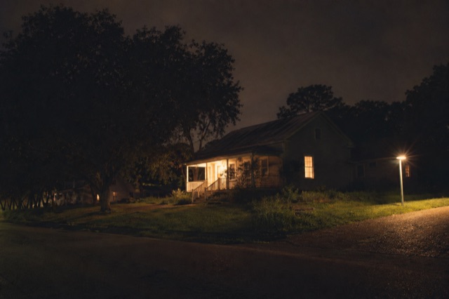 A Christian home at 11 PM, warm porch light, one window softly lit, a subtle extra light coming on at the corner of the property.
